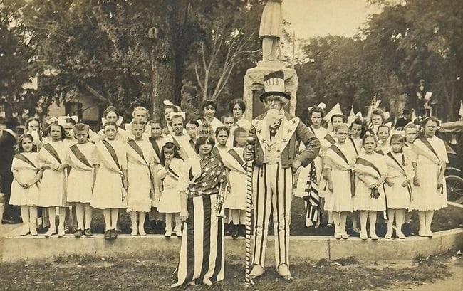 Historical Uncle Sam RPPC Patriotic Real Photo Postcard Parade American Flags: Historical patriotic real photo postcard featuring a man dressed as Uncle Sam with a young woman dressed as Lady Liberty or Miss Columbia. There are young girls with sashes in a town square. I do have