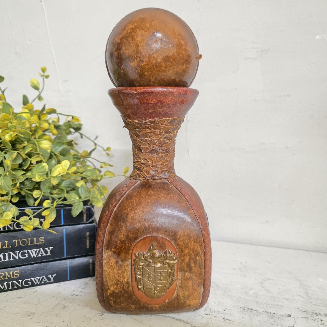 1970s Italian Leather Wrapped Decanter Brass Knight Crest, Bar Cart Dark Academic: This is a 1970s genuine leather-wrapped glass decanter made in Italy — distinguished by an applied brass heraldic coat of arms medallion on the front body, a knight's helmet crest above a