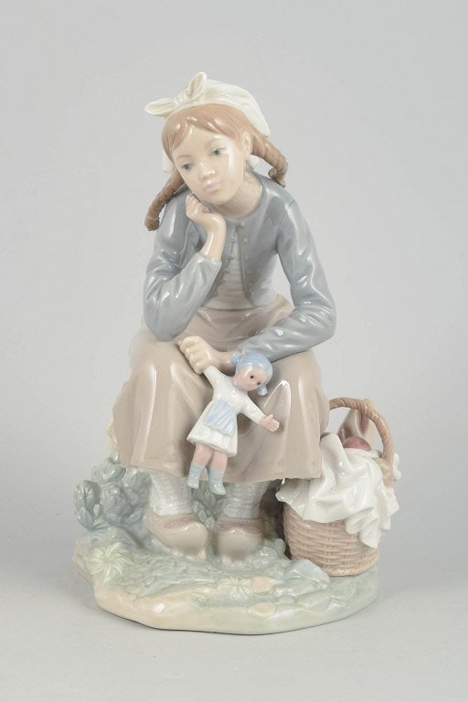 Lladró,, Spain. Porcelain figurine of a seated girl with doll in pastel tones. Approx. 1980s.: Lladró, figurine, seated girl with doll. Fine porcelain figurine depicting a contemplative girl seated with a doll at her side and a basket. Executed in soft pastel tones with characteristic glaz