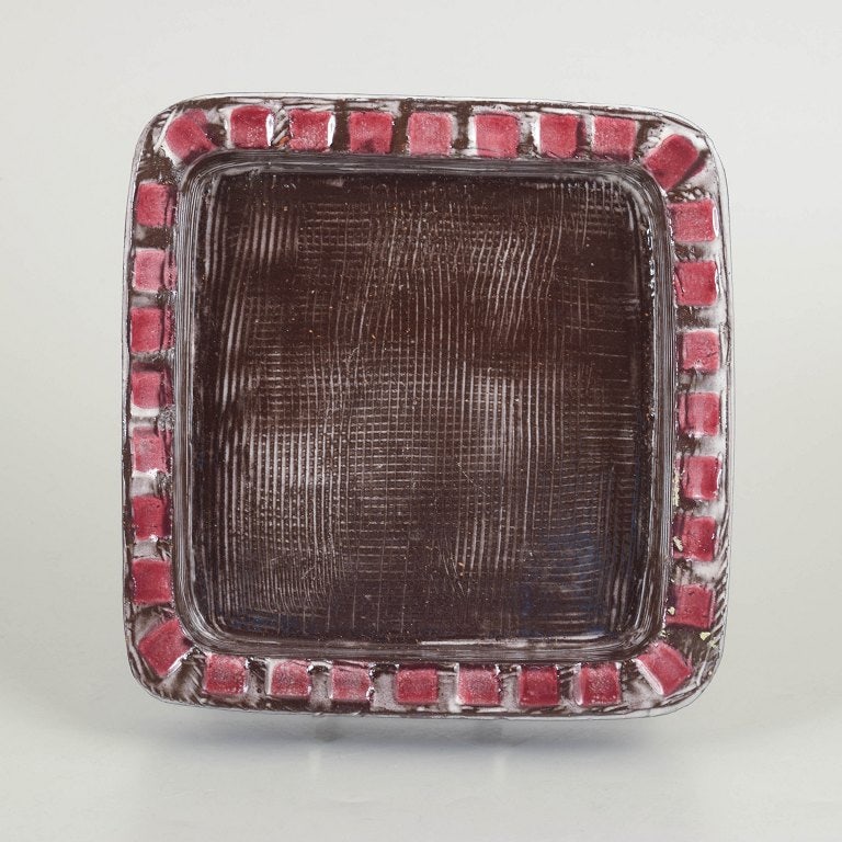 Mari Simmulson for Upsala Ekeby, Sweden. Stoneware dish with brown glazed centre. Approx. 1960s.: Mari Simmulson for Upsala Ekeby, dish. Square stoneware dish with a dark brown glazed centre and a decorative rim featuring relief and repeating red elements. Slightly textured surface. Model 4156. Or