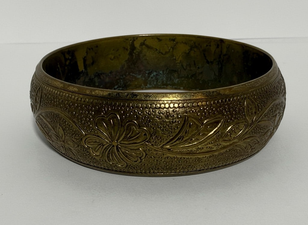 Vintage Embossed Brass Bangle Bracelet: This vintage brass bangle bracelet features an embossed floral and foliate design with a textured background. The bracelet has a concave interior profile and shows signs of age-related patina.