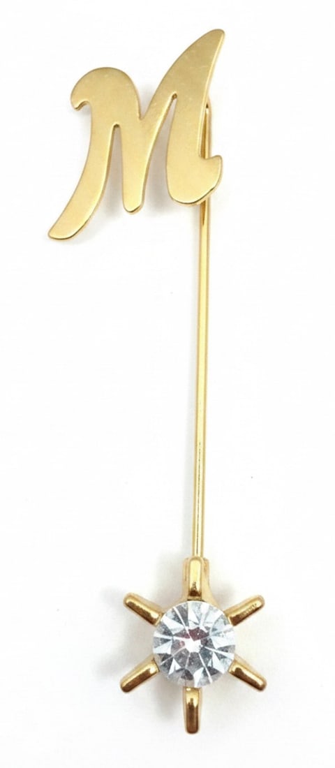 Gold-tone "M" Initial Stick Pin with Rhinestone Accent: A gold-tone stick pin featuring the initial "M" in a script font, accented with a single faceted clear rhinestone at the bottom. The rhinestone is set in a six-prong open-back setting. The pin has a