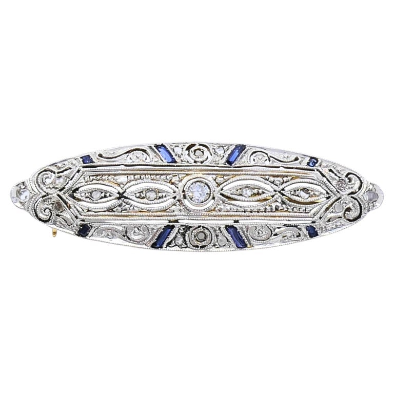 Art Deco 18k Gold Diamond Sapphire Navette Brooch: An Art Deco brooch in 18k gold, set with diamonds and blue sapphires in an elongated navette shape. The surface shows open milgrain filigree work with a central diamond flanked by smaller stones