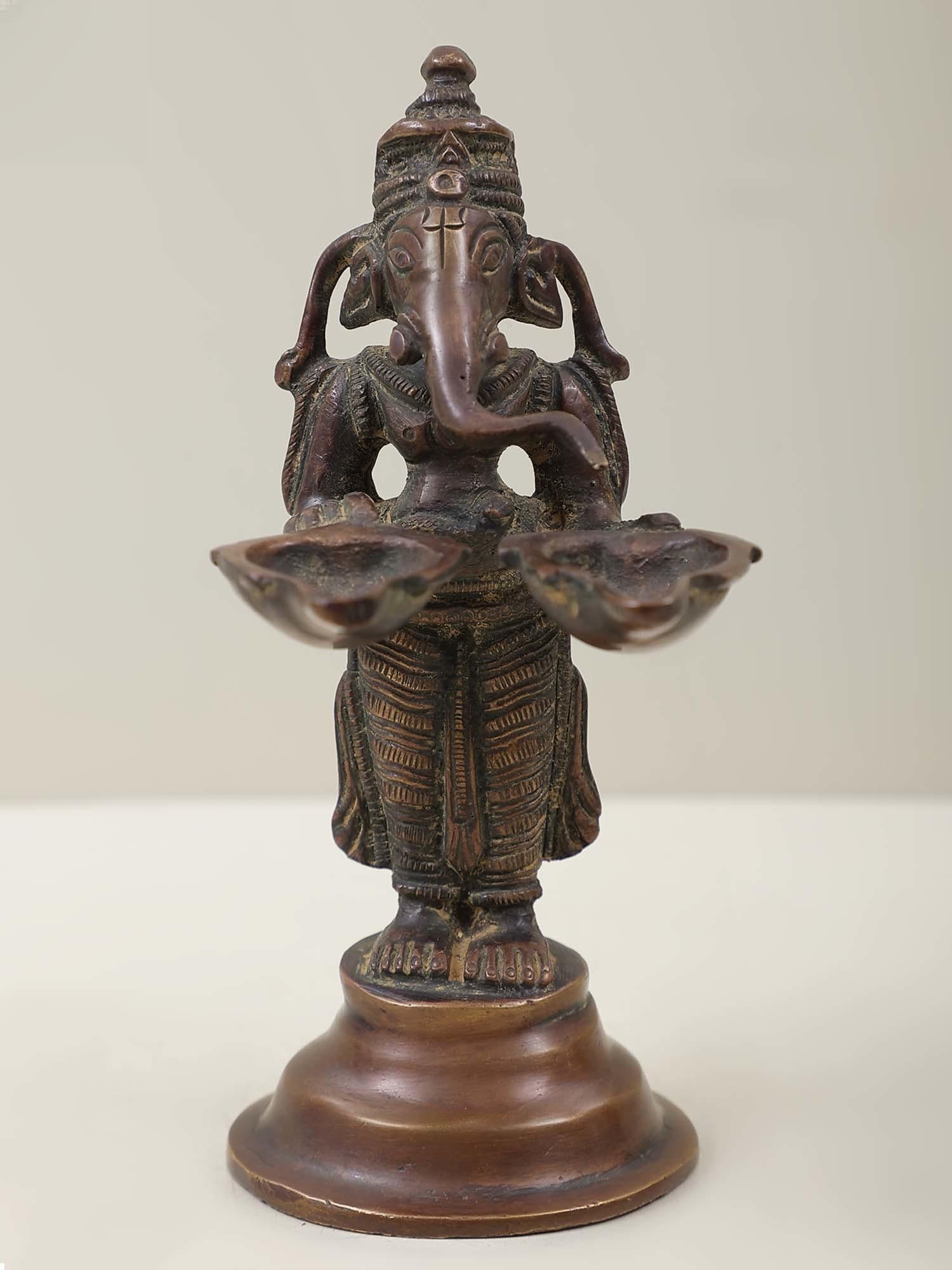 5" Lord Ganesha Twin Lamps In Brass | Handmade | Made In India: Description: See It In Your Home Light the lamps of wisdom within. In this exquisite brass statue , Lord Ganesha stands gracefully, flanked by twin lamps that illuminate the path of knowledge. As the