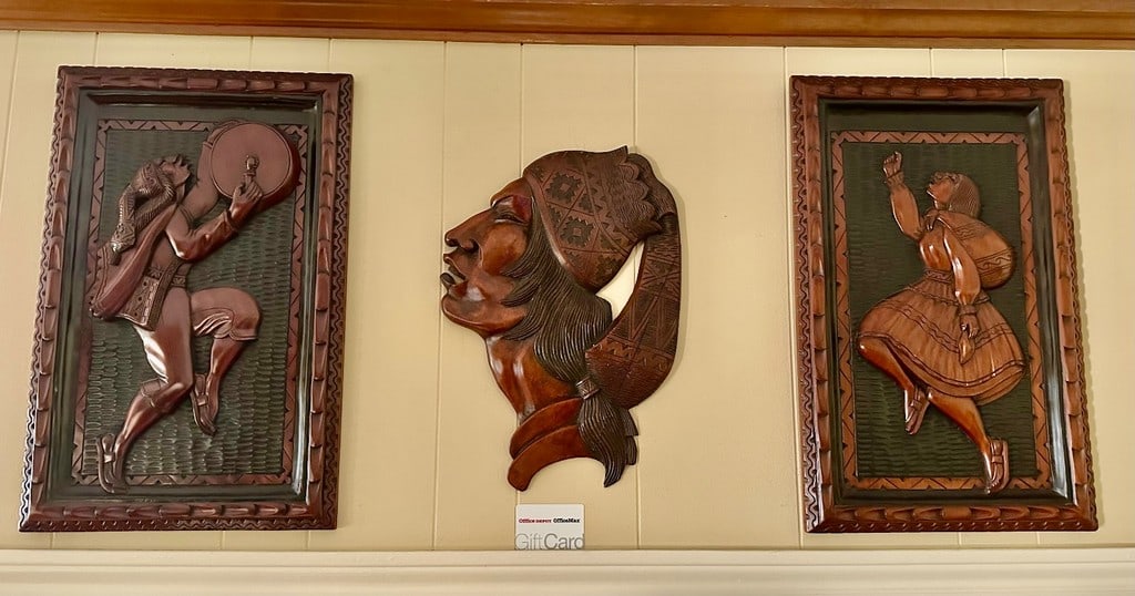 Set of Three Hand-Carved Bolivian Wood Wall Hangings: A set of three hand-carved wood wall hangings from Bolivia. The central piece depicts the profile of an indigenous person with intricate carvings on their headwear. The two framed pieces show figures