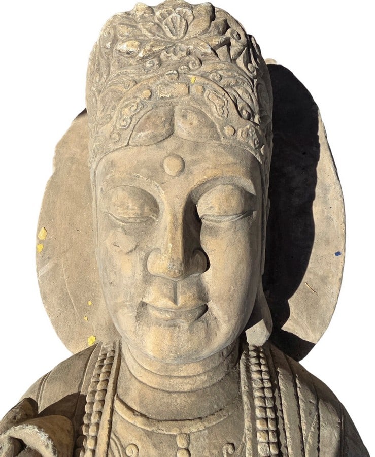 Carved Stone Buddha Bust: A serene carved stone bust depicting Buddha, featuring intricate detailing on the robes and headdress. The piece exhibits a weathered patina consistent with age and outdoor display, highlighting the