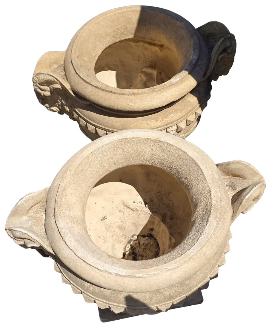 Pair of Cast Stone Neoclassical Style Garden Urns: A pair of weathered cast stone garden urns, each featuring a wide, ribbed rim over a classical body adorned with ram's heads and a lower register of acanthus-like detailing. The urns exhibit a patinat