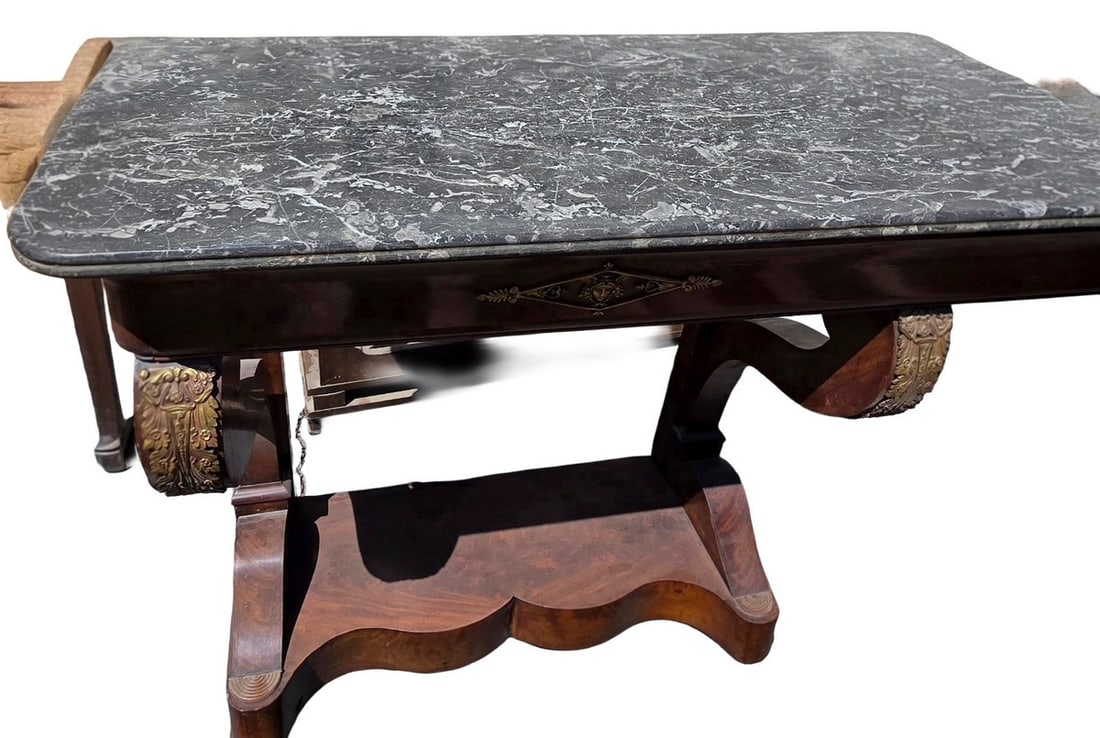 Antique Empire Style Marble Top Console Table: An antique Empire style console table featuring a rectangular grey marble top over a richly veneered mahogany base. The base is characterized by scrolling supports, decorative bronze ormolu mounts, an
