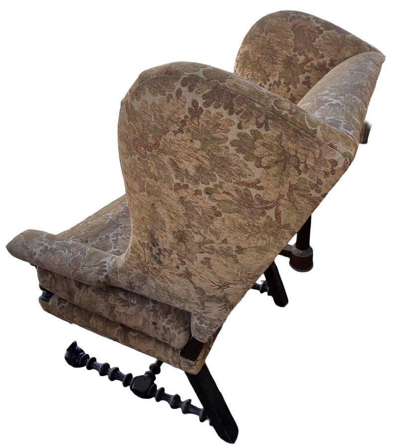 Vintage Upholstered Wingback Armchair with Carved Wood Frame: A vintage wingback armchair featuring an upholstered frame with a botanical pattern in shades of brown and green. The chair has a distinctive carved wood base and legs with turned details, suggesting 