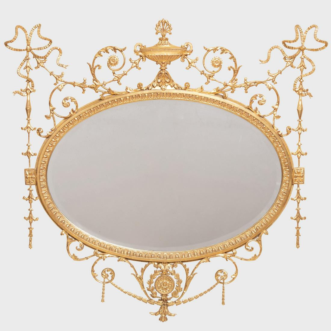 George III Style Giltwood Oval Mirror: George III Style Giltwood Oval Mirror Fitted with a beveled mirror plate, surmounted by a classical urn issuing ribbon-tied foliate scrolls, terminating in a pierced medallion. 47 x 48 3/4 in.