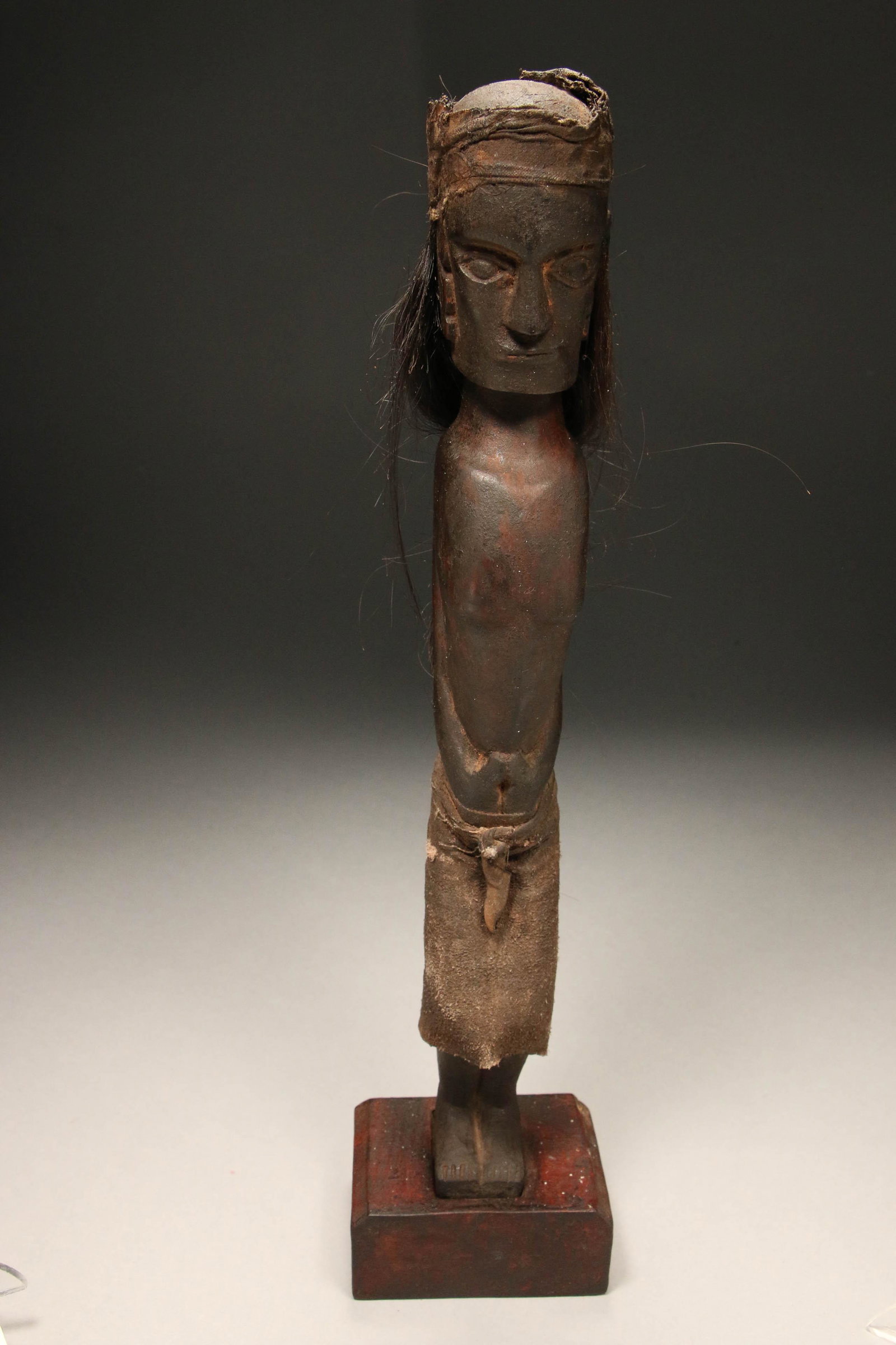Indonesia Batak Statue: A powerful Batak ancestor statue with human hair, cloth head band and sarong. Cord belt with fabric container. On wood block stand. 16 H " (w/stand)