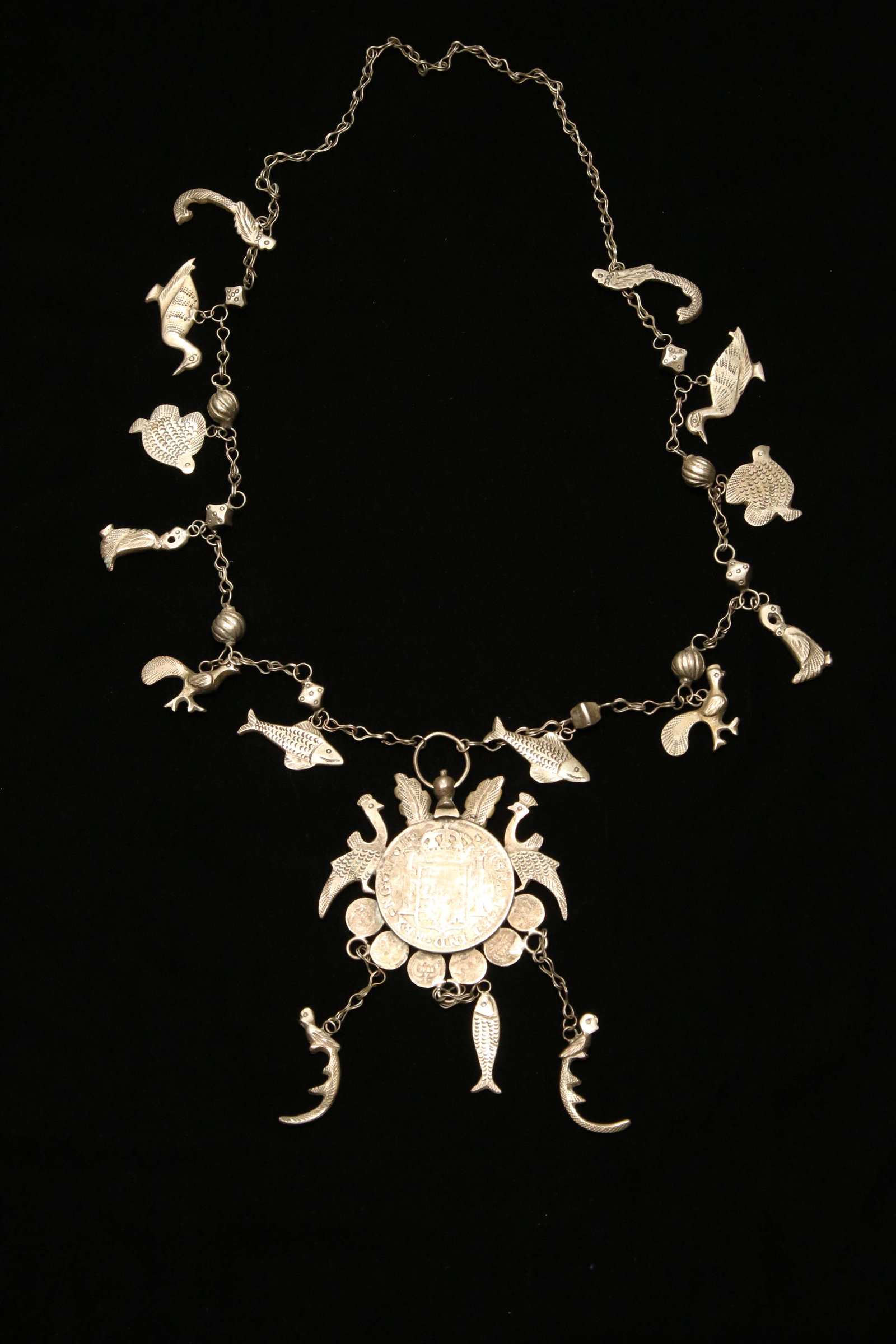 Antique Silver Coin Prosperity Necklace: A silver necklace with a Spanish Ferdin 1818 coin fashioned into a central pendant. The necklace is surrounded with silver animals, peacocks and fish hanging from double link chains. The coin is surro