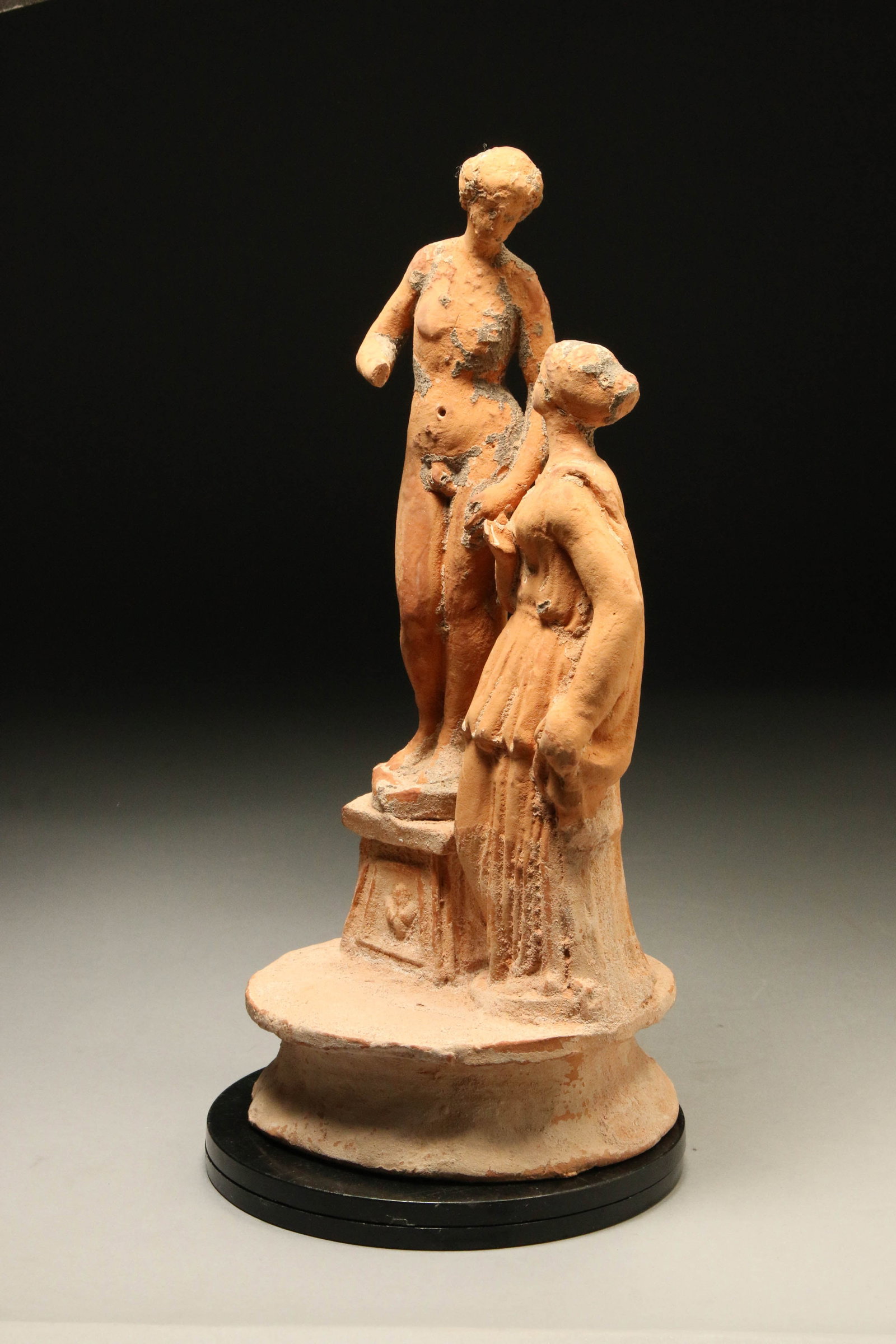 Ancient Greek Terracotta Statue - Two Figures: A large ancient Greek pair statue. A nude Adonis, his right leg slightly bent on the knee, standing on a raised pedestal, gazing down on an enamoring Aphrodite, with hair pulled back into a bun, her r