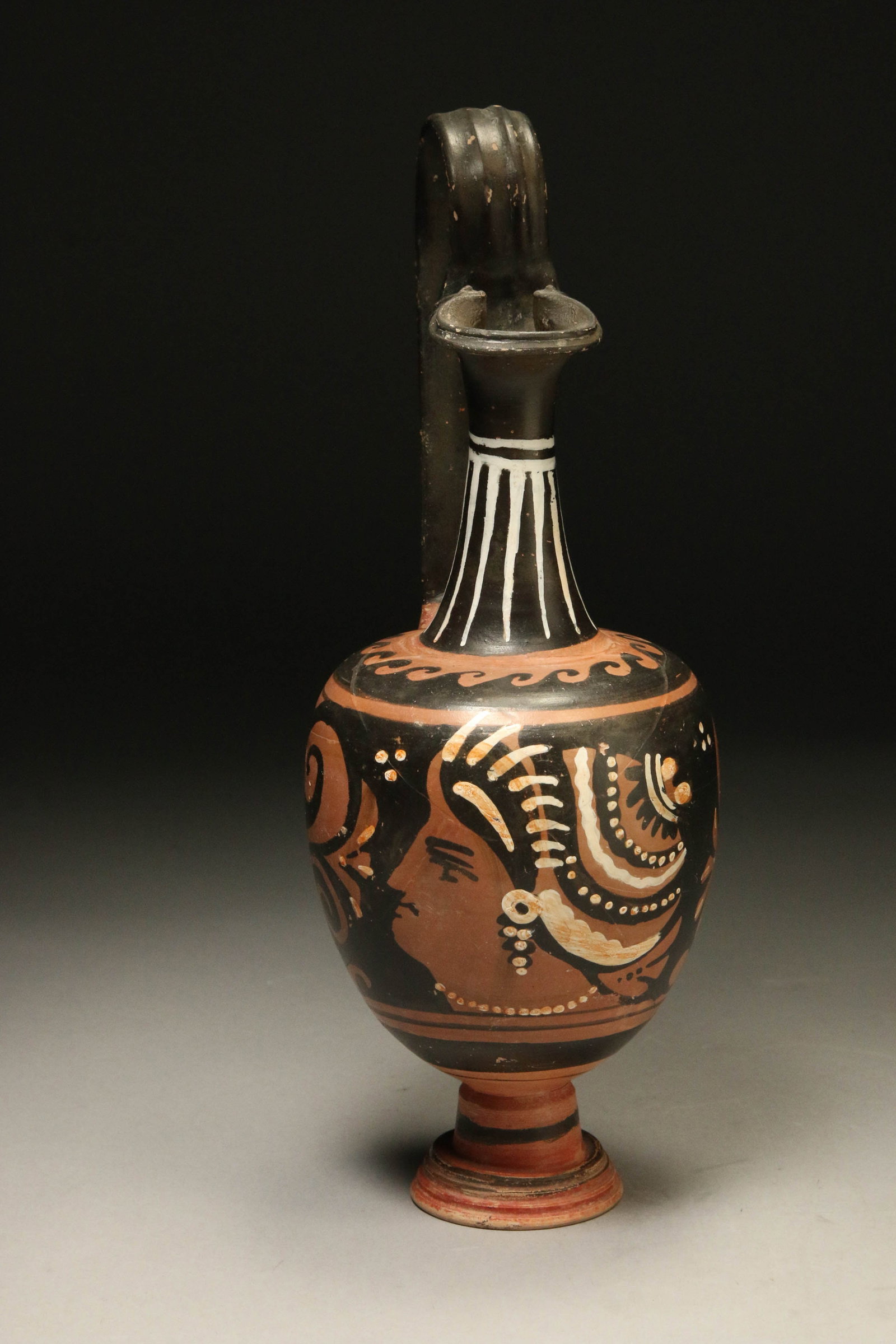 Ancient Greek Oinochoe, ca. 325 - 300 B.C.: A Greek terracotta Oinochoe with painted female profile. Strap handle, fluted neck with trefoil mouth. Seating on a flared, pedestal base. 11" H Provenance: Ex collection New York City.
