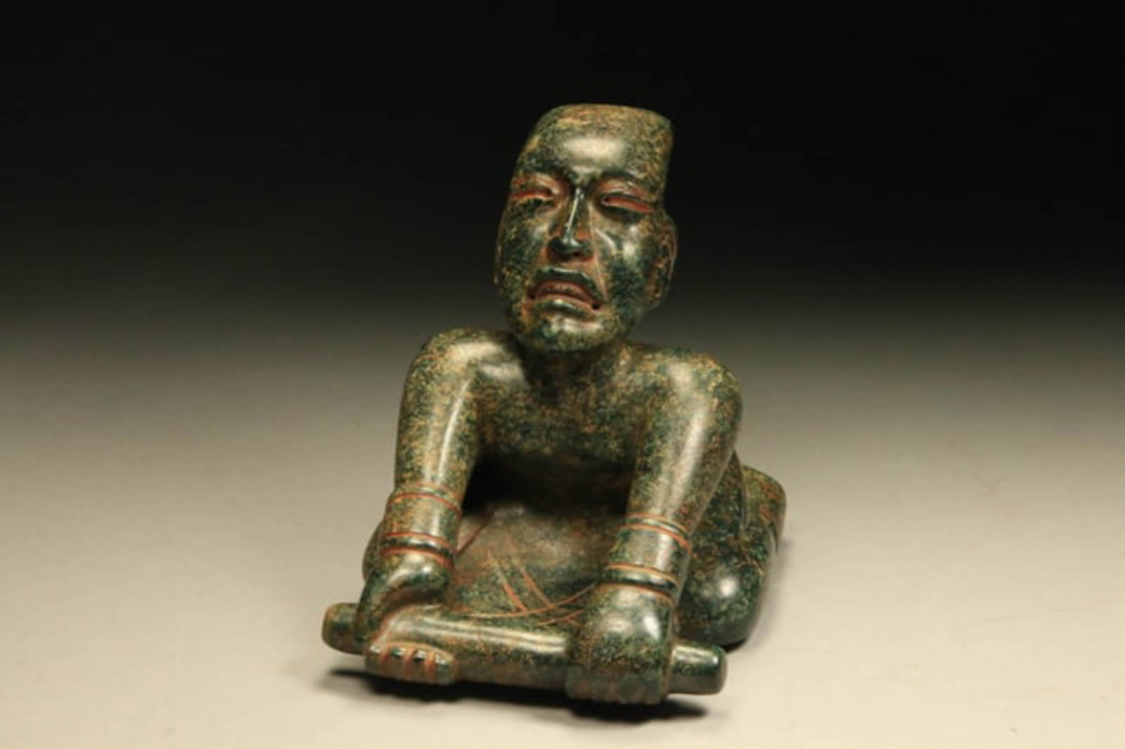 Pre-Columbian Olmec Figure: A museum quality carved jade figure of a kneeling figure holding a scepter. Classic Olmec ovoid head with open downturned mouth. Incised designs throughout. Back depicts a protruding spine and figure 