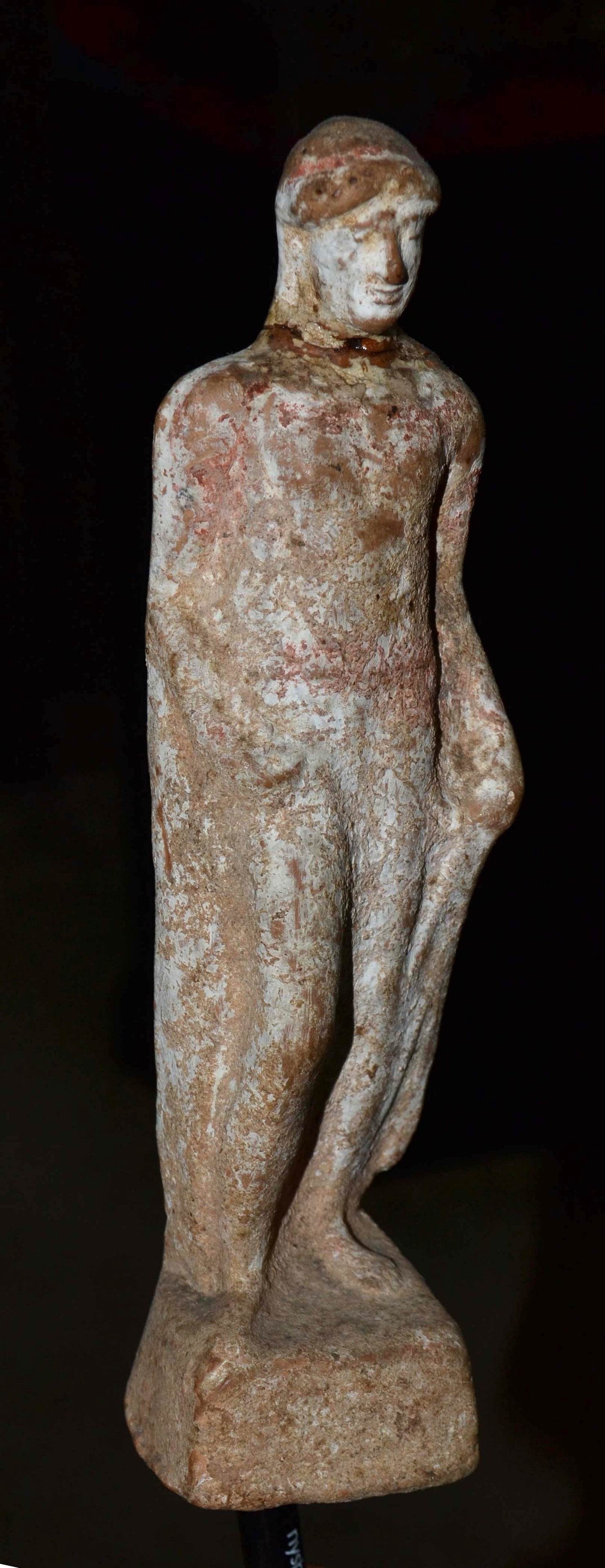 Ancient Greek Terracotta: A Greek terracotta of a young man. With defined facial details, wearing a covering over his head, standing on a pedestal base, right leg bent on the knee, wearing a long drapery, falling down his back