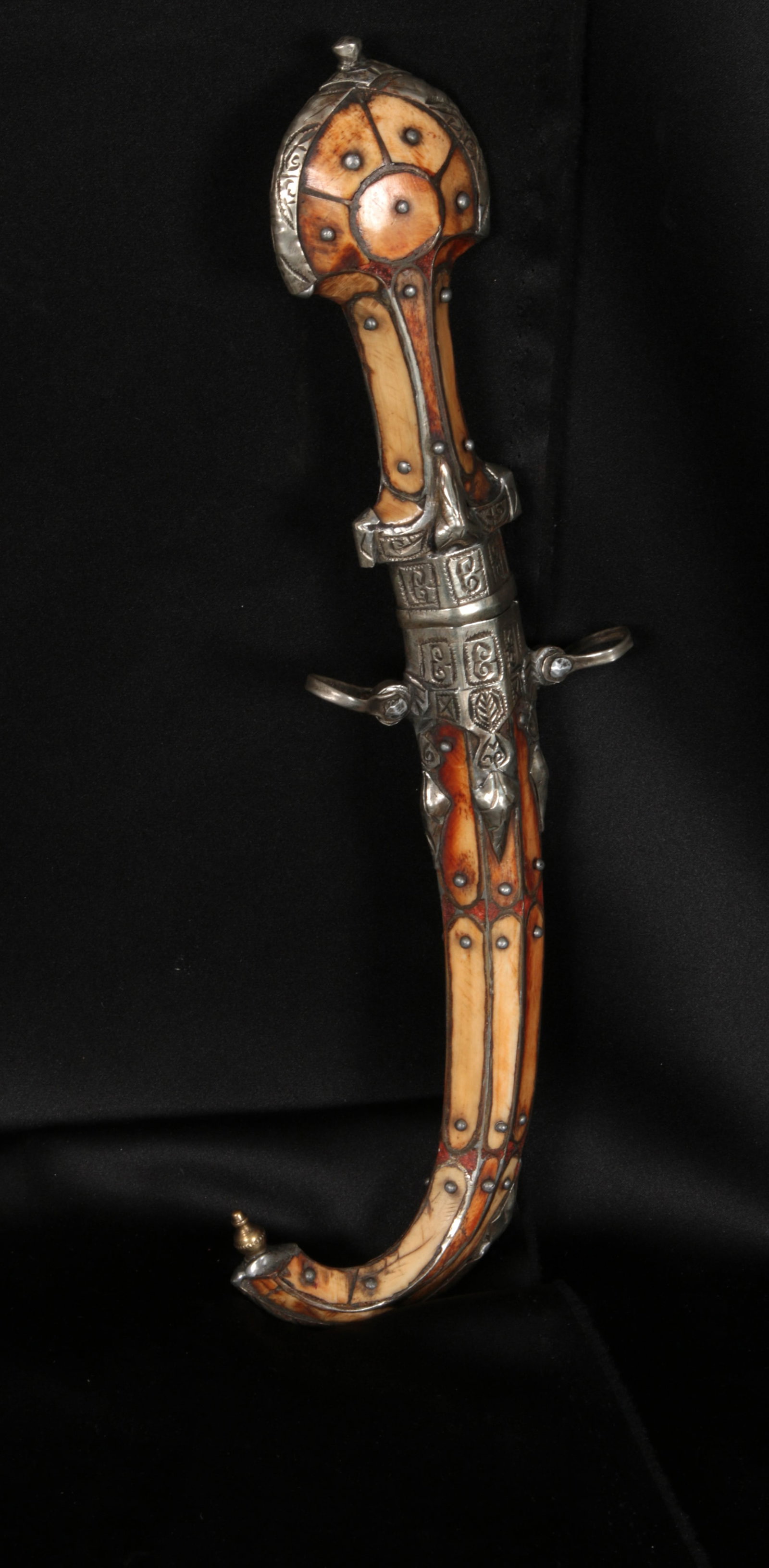 Moroccan Koummya Dagger: A curved dagger made of bone, silver and mixed metal. The hilt has a stylized pea-cock tail shaped pommel, the scabbard has silver trim, suspension loops and a metal tip. A curved steel blade is house