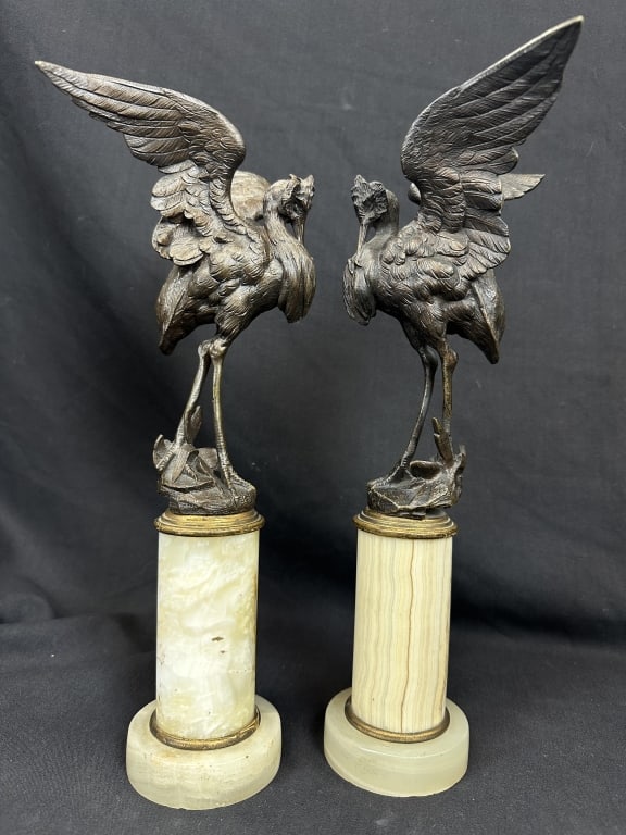 Pair of Bronze Heron Figures on Marble Columns: A decorative pair of patinated bronze figures depicting herons in animated postures, each bird shown poised after landing with wings partially extended. The sculptural treatment emphasizes movement an