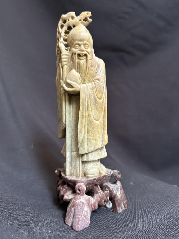 Chinese Carved Soapstone Shoulao Figure: A Chinese carved soapstone figure depicting Shoulao, the Daoist god of longevity, modeled as a bearded elderly sage holding a staff. The pale stone figure is mounted on a contrasting reddish-brown car