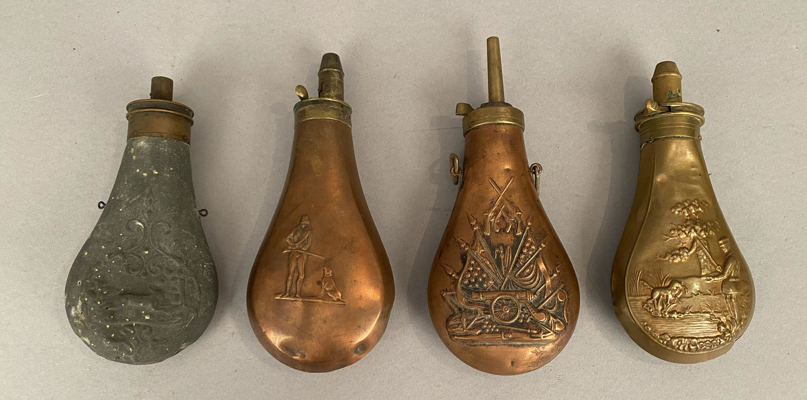 4 Antique Black Powder Flasks w/Embossed Designs: Four antique black powder flasks, all with figural Embossed designs. One with a cannon, flags and guns, 2 with a man and dog and one with a rabbit. Made of copper or brass. Largest 8"