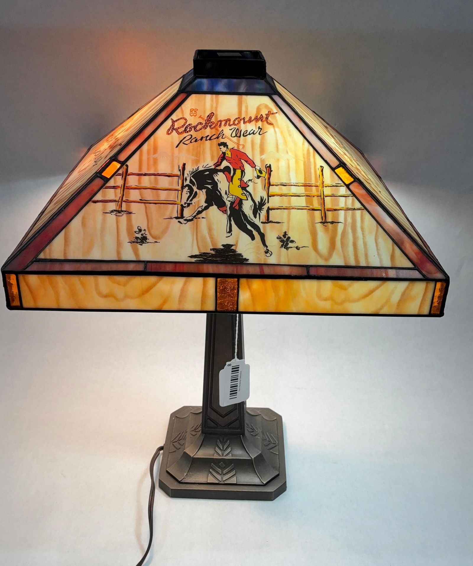 Rockmount Ranchwear Hand Painted Western Stained Glass Lamp: Showing a cowboy to two panes and Rockmount logo to all sides. No cracks or chips found. Shade is 16x16. A heavy art deco base includes two bulbs. Entire height is 23 inches.