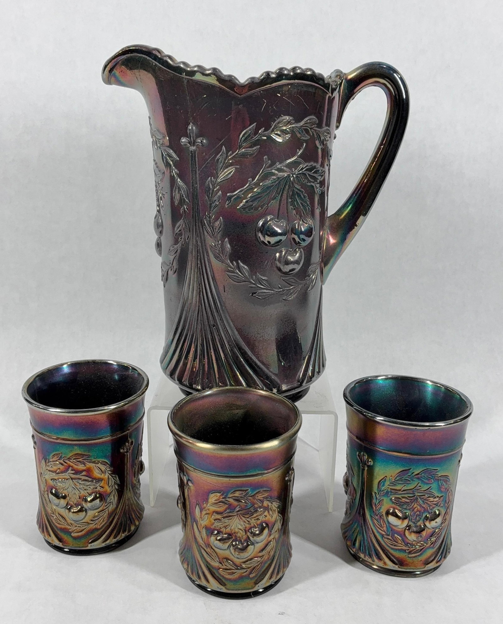 Dugan Cherries in Wreath Water Pitcher & Tumblers: Ca 1901-1915, 9 inch tall pitcher with three matching glasses. One cup shows a small chip to bottom and pitcher has a flea bite to detailed side. Rainbow like sheen to all pieces with a deep purple
