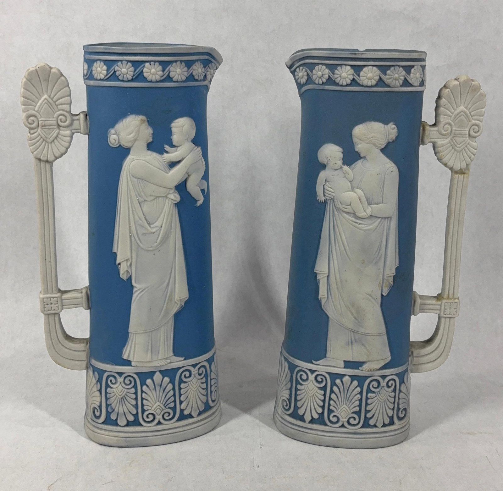 Jasperware Mother & Child Neoclassical Style Pitchers: In the Style of Wedgewood but not marked. Each pitcher shows a mother and child with decorated handle and pattern along top and bottom. Each is 8.5 inches. One shows a few small chips from rim.