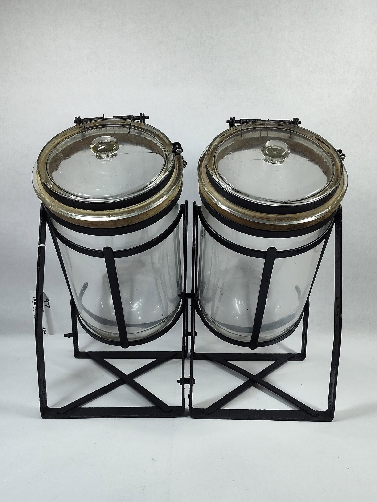 Heavy Glass Country Store or Medical Storage Jars: Heavy glass containers approximately 2 gallon capacity with iron banded hinged lids and rounded bottoms in wrought iron frame. One of the lids has a chip, otherwise they appear good. Unit is 16.5in