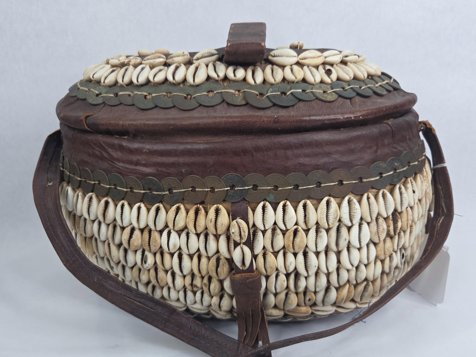 Handmade Cowrie Shell Leather Hausa Basket: Large handcrafted straw basket with leather made in Nigeria by the Hausa people. Adorned with Nigerian tokens around lid and mouth of basket and numerous Cowrie shells. 14in long 10in wide 10in tall.