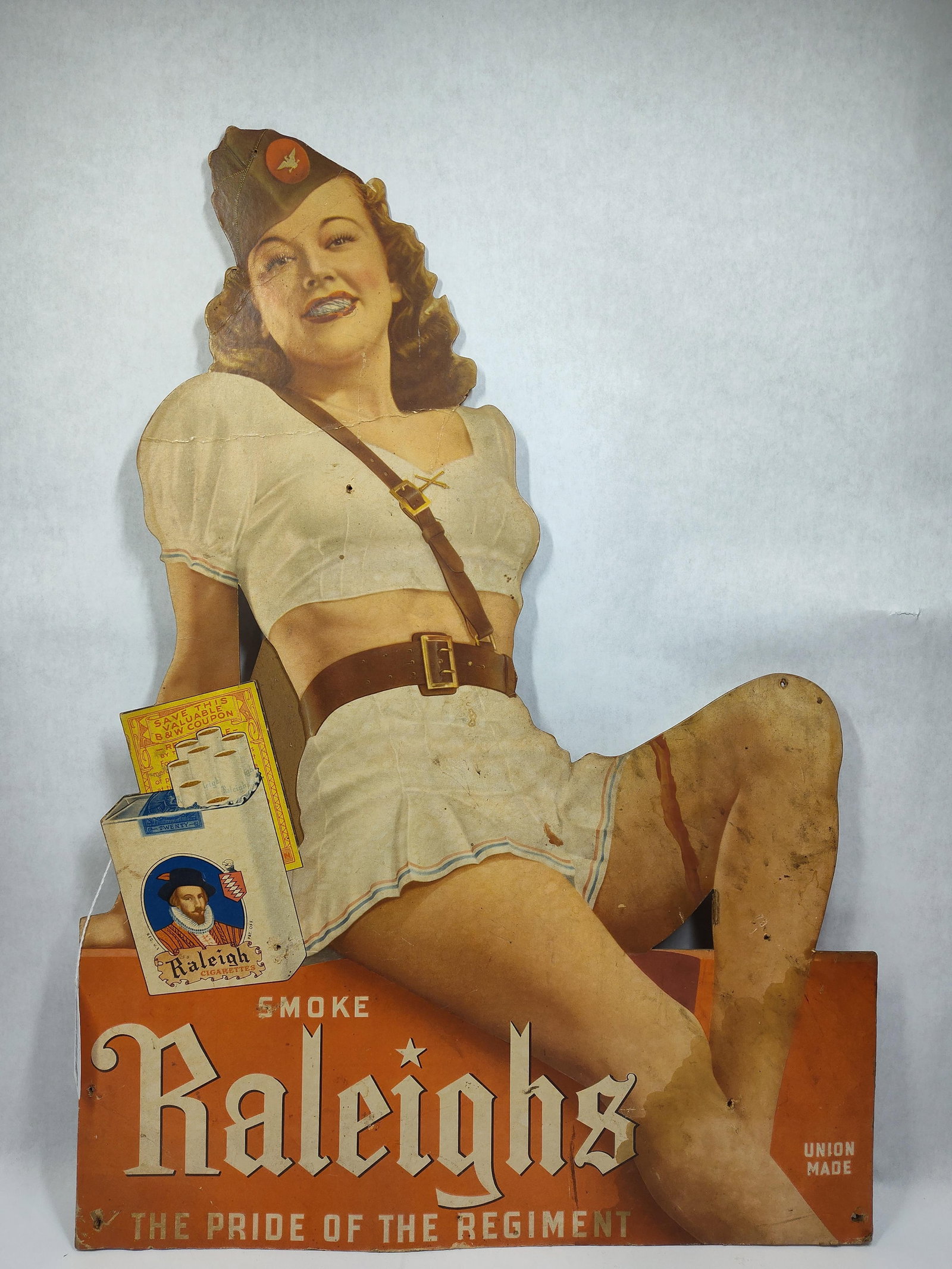 Raleighs Cigarettes cdbd easelback sign: 1-sided cardboard litho easelback sign with some soiling, edge wear and a few nail holes. Also shows a paint run on leg, skirt and pen scribbles on teeth but is a nice graphical piece! 20in x 28in.