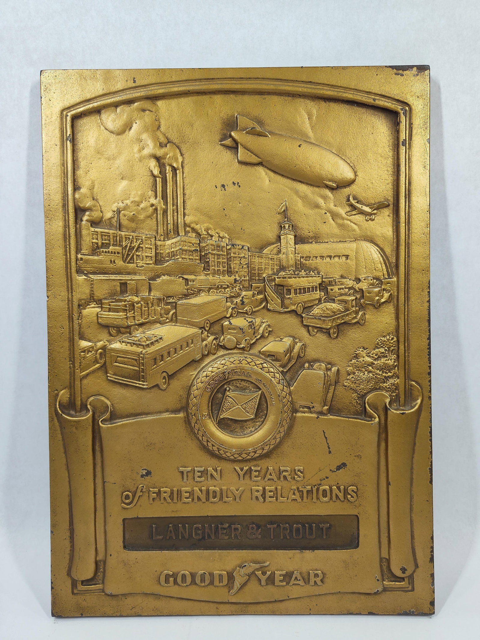 Great Goodyear 10 yrs Friendly Relations Dealer Plaque: Cast metal/Bronze embossed plaque depicting Goodyear factory, early cars, trucks, train, airplane and blimp. Wonderful example with minor edge wear but overall in great condition! Marked Medallic Art