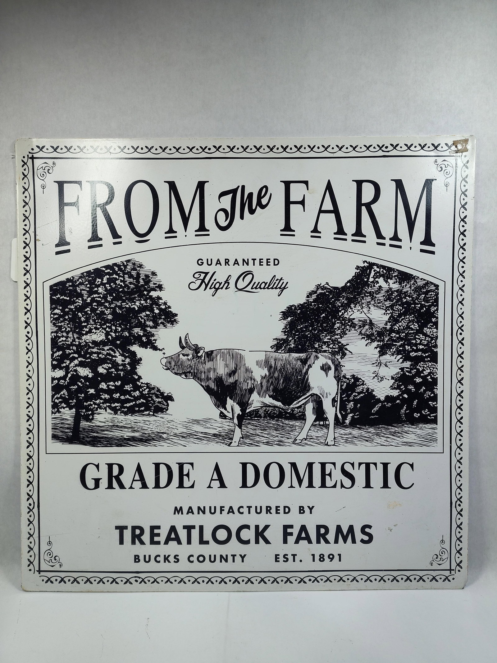 Decorative Metal Farmhouse Wall Sign featuring Cow Illustration and Treatlock Farms Branding: This decorative metal wall sign features a black and white graphic design in afarmhouse style. The central illustration depicts a cow standing in a field between two deciduous trees. The piece is