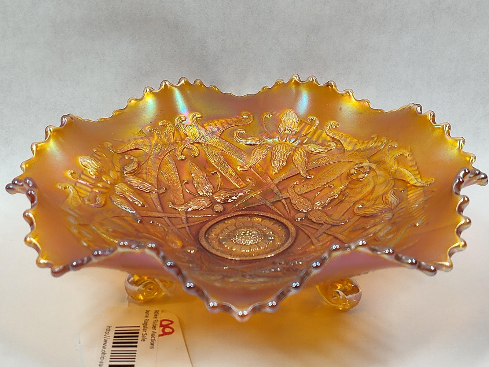 Marigold Carnival Glass Footed Bowl in the Iris Pattern: This tri-footed bowl is a specimen of American carnival glass, featuring a marigold iridescent finish that exhibits varied lusters of gold, pink, and blue. The interior surface is decorated with a pre