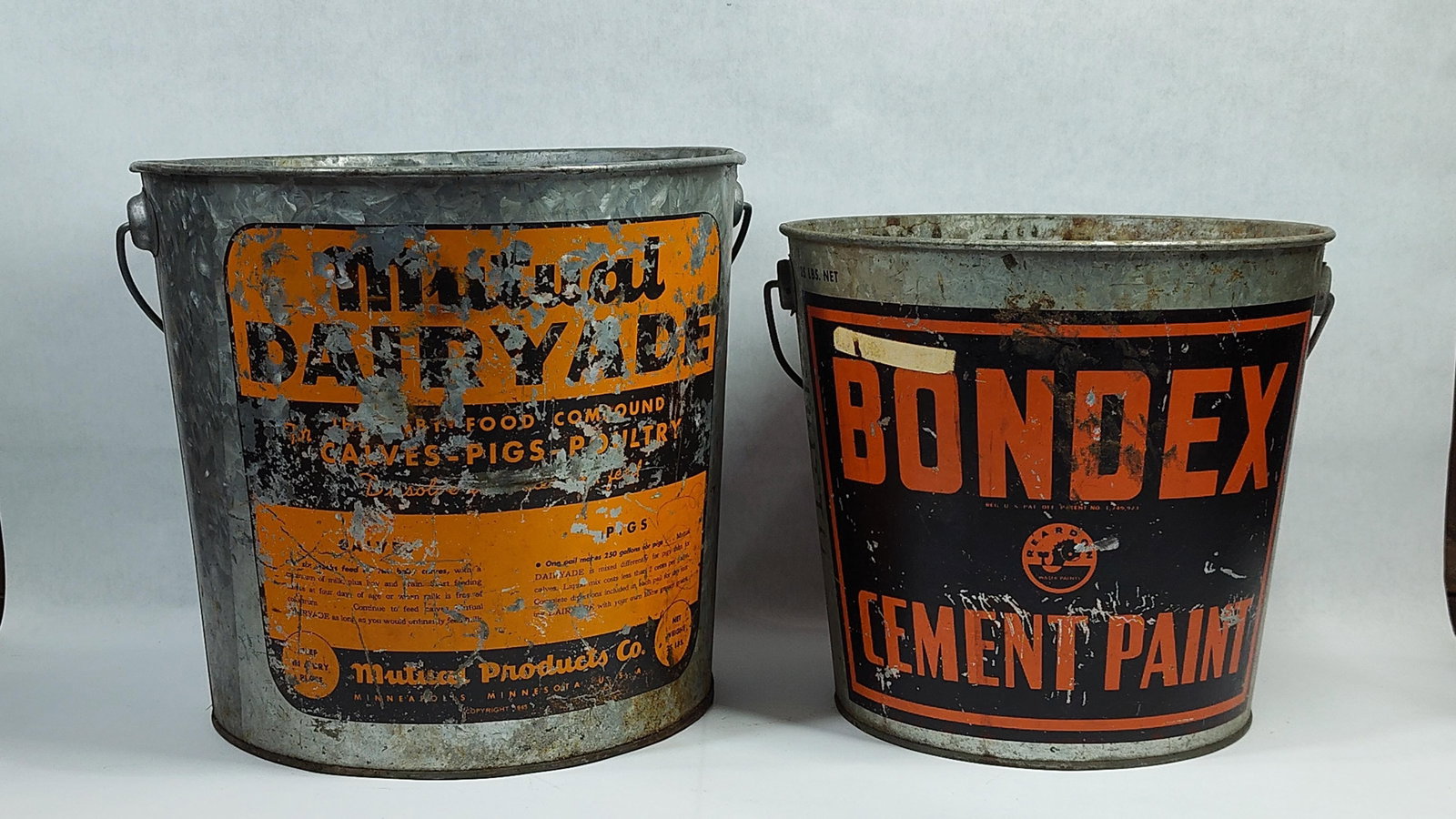 A Pair ofGalvanized Metal Advertising Buckets for Bondex and Mutual Dairyade: This lot consists of two mid-20th-century galvanized steel advertising pails, each equipped with its original wire bail handle. The first bucket features an orange and black label for Mutual