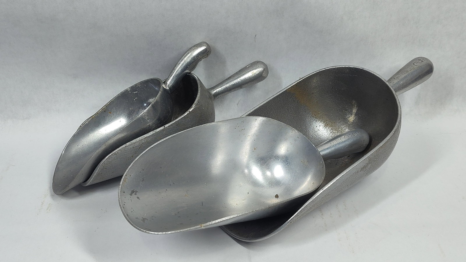 A Set of Cast Aluminum Commercial Utility Scoops: This lot includes four graduated utility scoops constructed from cast aluminum, intended for commercial kitchen or dry goods applications. One scoop is marked WINCO 24OZ on its handle. A second, small