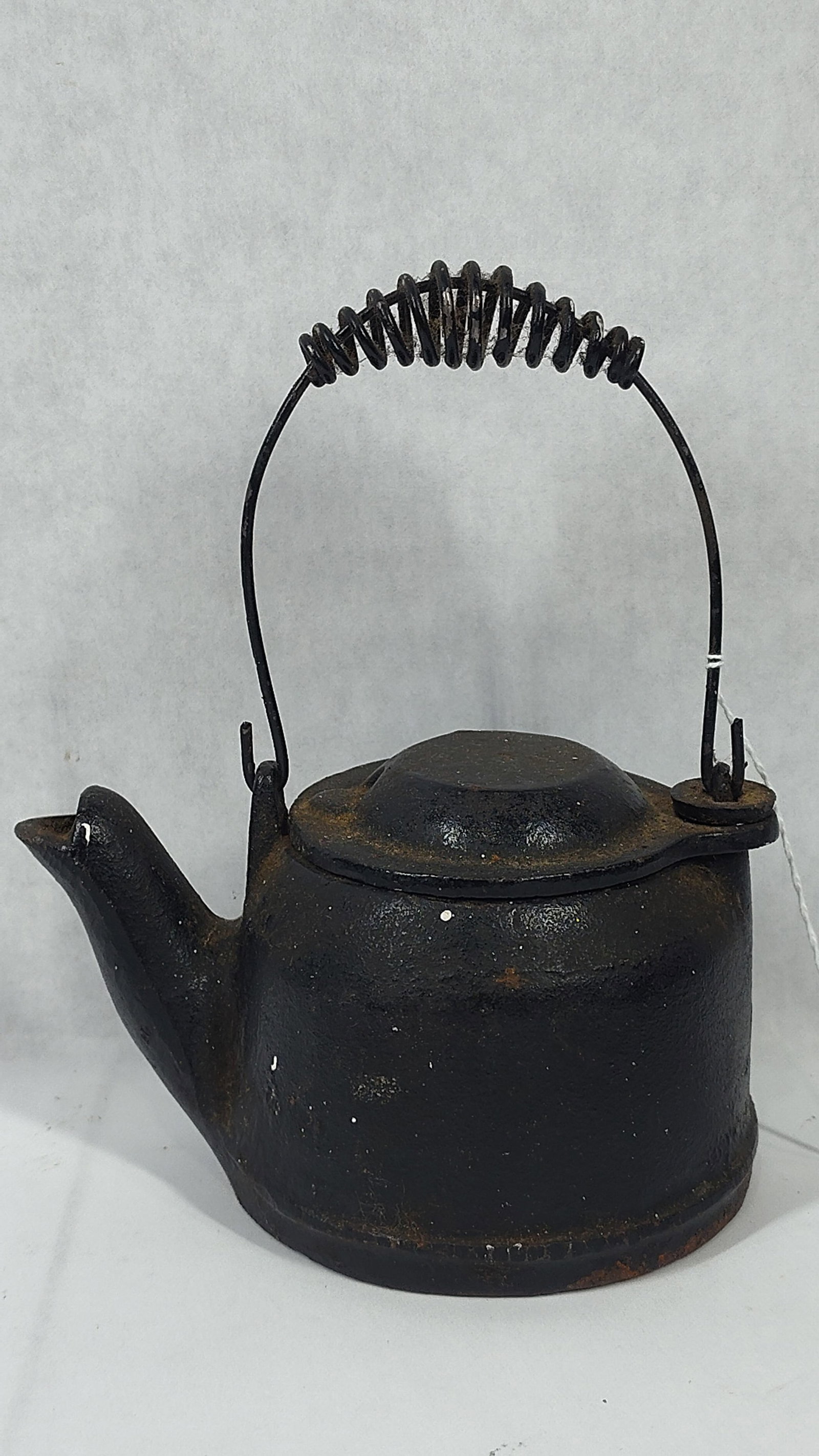 Cast Iron Tea Kettle with Swivel Lid and Coil Wire Bail Handle: A small cast iron kettle constructed with a squat, bulbous body and a short, integral spout. The vessel features a swivel-style lid secured by a single-point hinge. It is equipped with a wire bail han