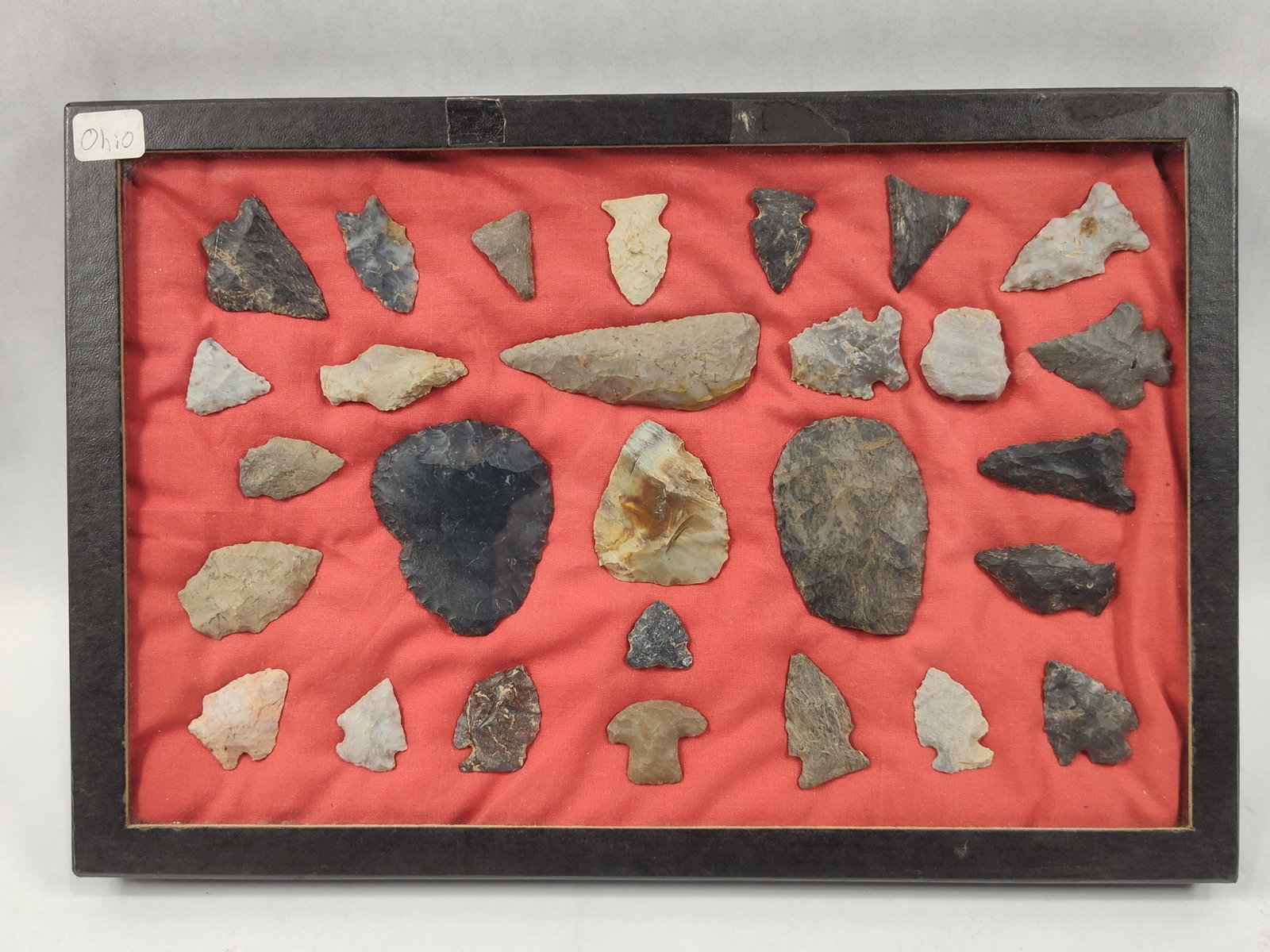 Native American Lithic Artifacts from Ohio: This is twenty-eight Native American lithic artifacts, primarily consisting of projectile points and bifaces. The group is presented within a glass-fronted display case with a red fabric lining.