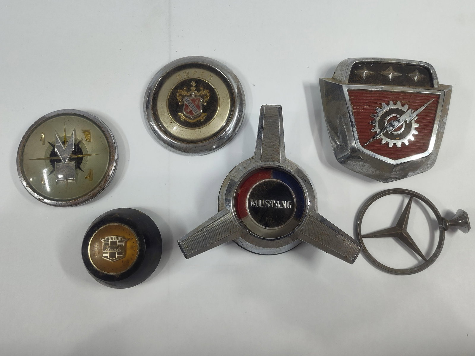 American and European Automotive Emblems and Ornaments: This collection contains severalmid-century automotive emblems, center caps, and ornaments from various manufacturers. The lot includes a chrome-plated tri-bar wheel spinner center cap featuring a
