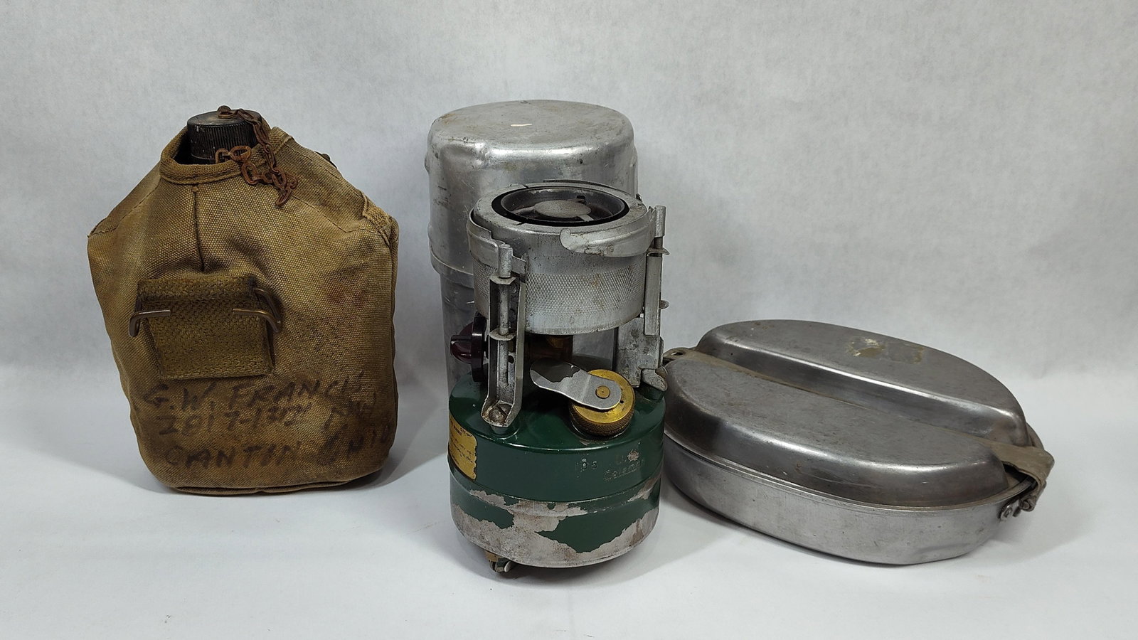 United States Military Field Gear Including M-1950 Stove, 1945-Dated Mess Kit, and Named Canteen: mid-20th century United States military field equipment comprising several individual components. The group features a Model M-1950 pressurized single-burner gasoline cooking stove. This unit consists
