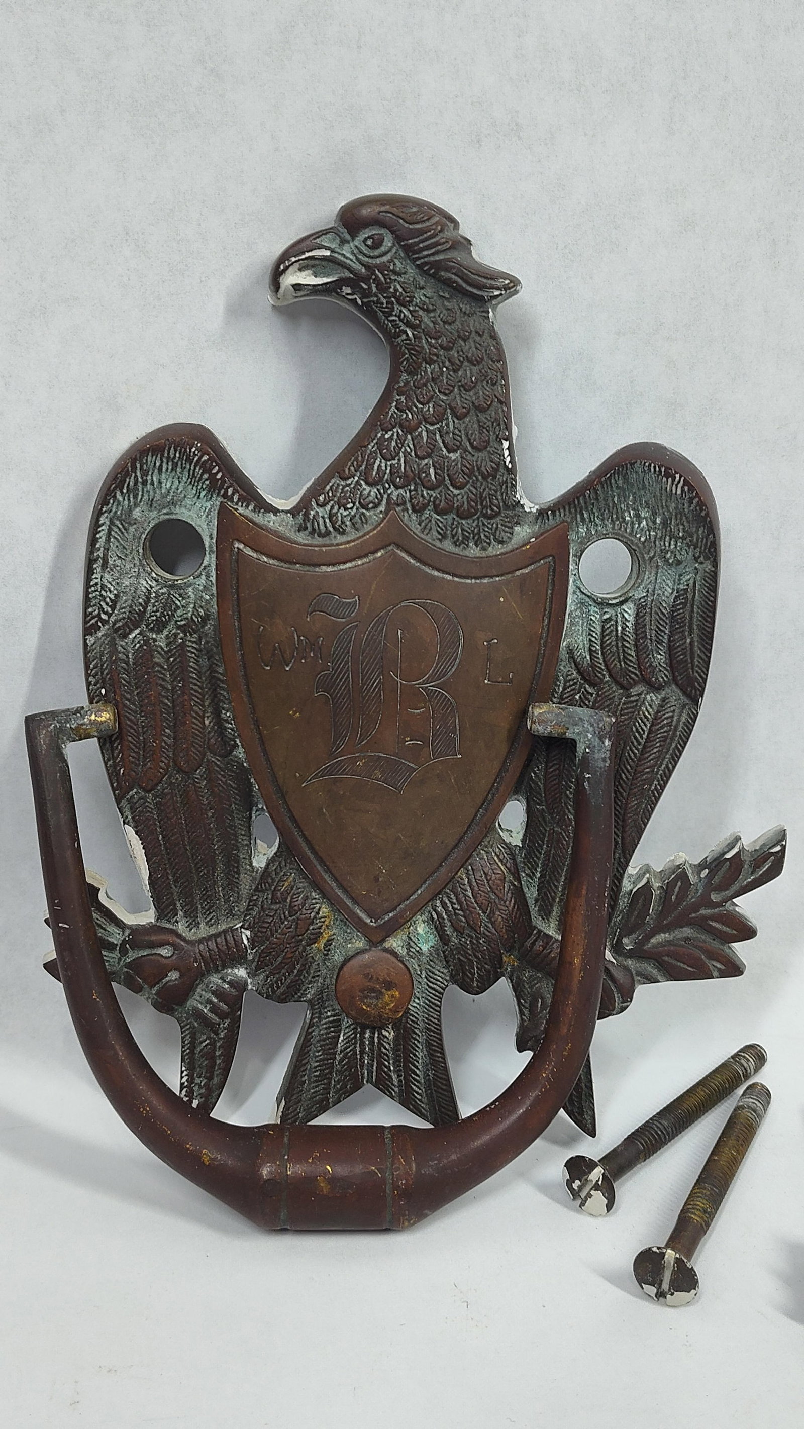 American Federal Style Cast Brass Eagle Door Knocker with Monogrammed Shield: Acast metal door knocker, likely brass or bronze, designed in the American Federal style. The backplate is formed as an eagle with outspread wings and its head turned to the dexter side. The eagle clu