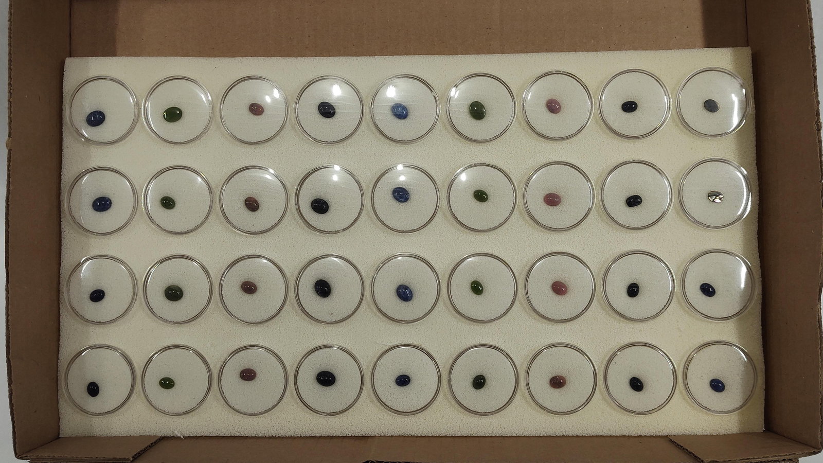 36 Loose Oval Gemstone Cabochons: This collection consists of 36 loose oval gemstone cabochons, each presented in an individual plastic gem jar with a white foam insert. The stones are cut in a uniform oval cabochon style with polishe