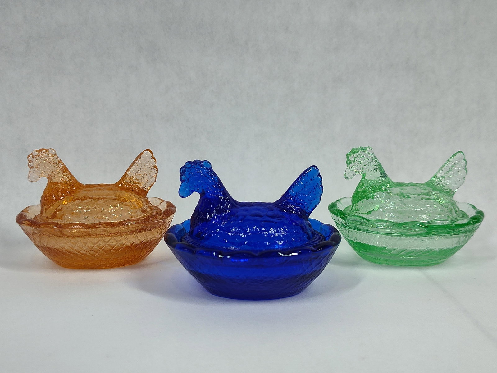 A Trio of Miniature Pressed Glass Hen on Nest Salt Cellars in Assorted Colors: This lot features three miniature two-part lidded dishes, traditionally used as individual salt cellars, designed in the form of a hen on a nest. The collection includes one cobalt blue, one amber, an