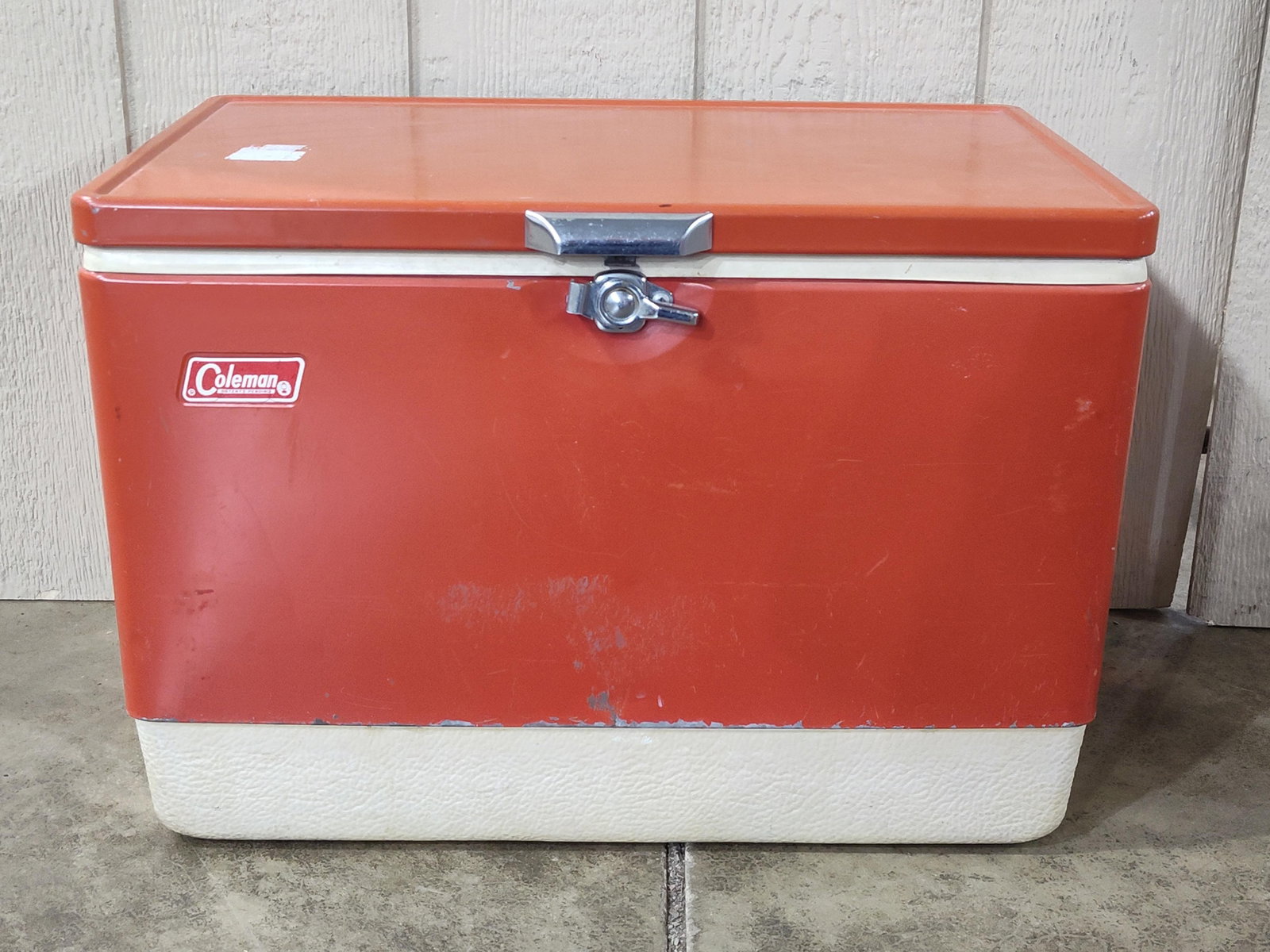 Coleman Red Metal and Plastic Portable Ice Chest Cooler: ThisColeman portable cooler features a two-tone construction consisting of an orange-red metal upper body and lid over a textured off-white plastic base. The front face of the metal body bears a red