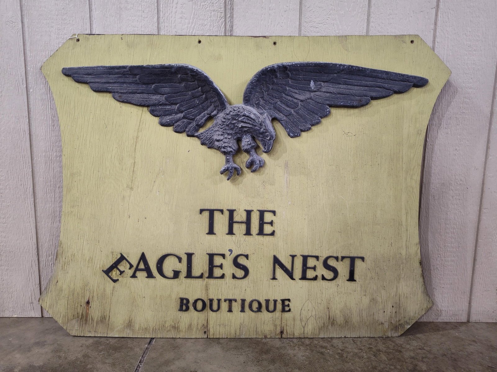 Hand-Painted Wood and Cast Metal Trade Sign for The Eagle's Nest Boutique: This is acommercial trade sign for a business titled The Eagle's Nest Boutique. The sign is constructed on a thick plywood panel featuring a shaped silhouette with curved sides. Mounted at the top is 