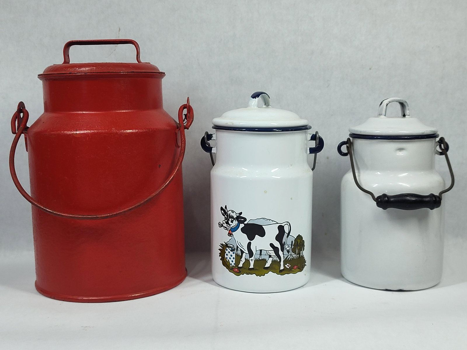Trio ofMetal and Enamelware Milk Cans: This lot comprises threemilk cans of varying sizes and finishes. The collection includes one large painted metal can, one medium enamelware can with graphic decoration, and one small plain enamelware 