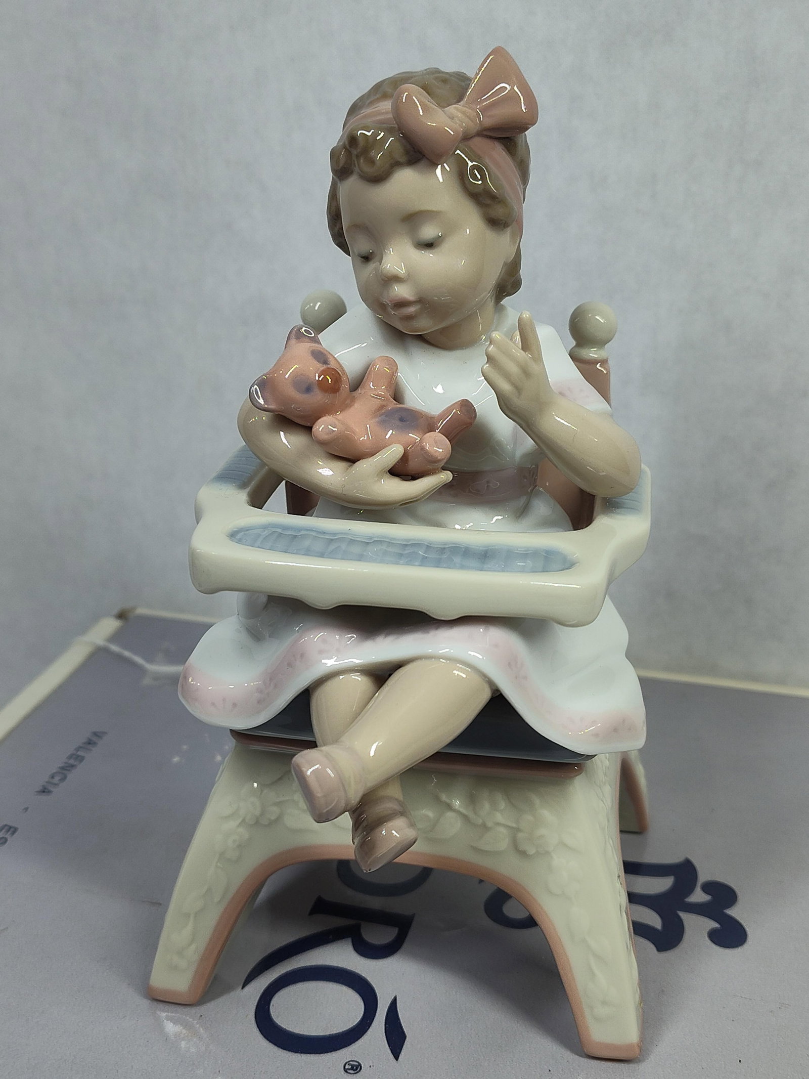 Lladro Porcelain Figurine Little Bear Model 6299: A glazed porcelain figurine produced by Lladro, titled Little Bear or Jugando con el osito, model number 6299. The piece depicts a young girl seated in a high chair, holding a small pink teddy bear. T