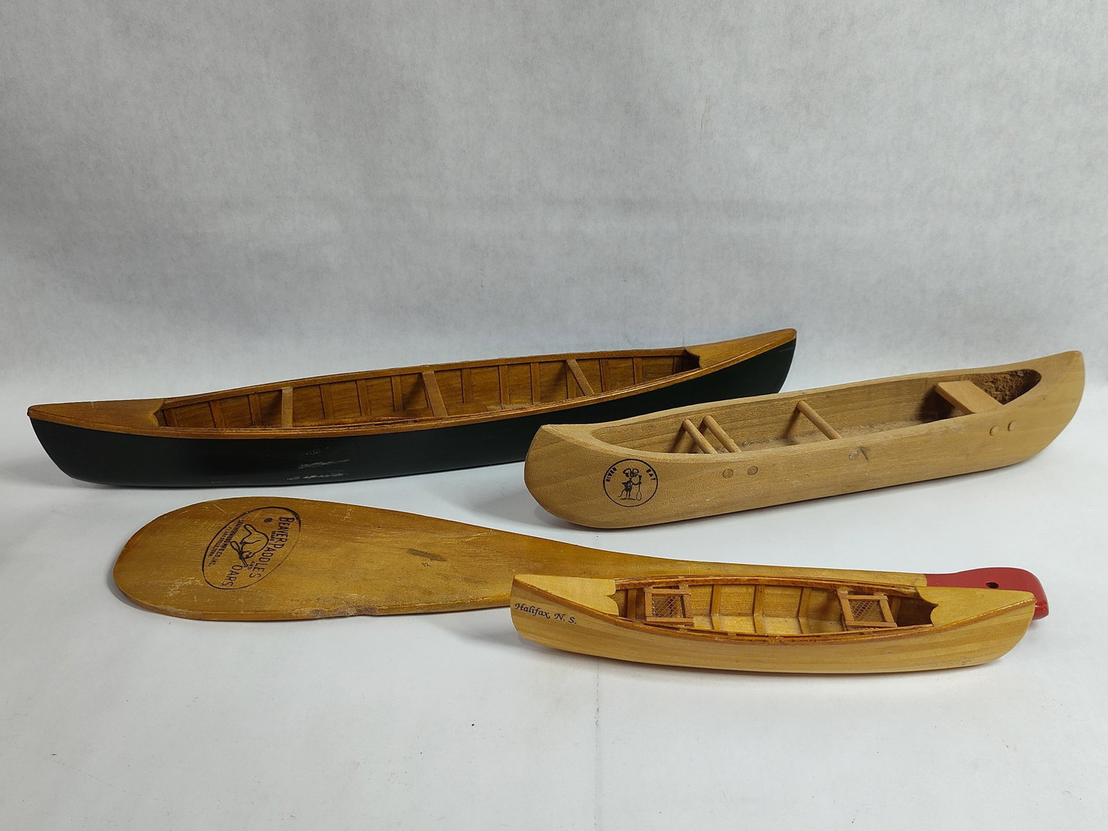 A Group of Wooden Model Canoes and a Decorative Beaver Brand Paddle: This lot comprises a group of three wooden model canoes and one wooden canoe paddle. One canoe features a dark green painted exterior hull and a natural wood interior with detailed ribbing and cross-t