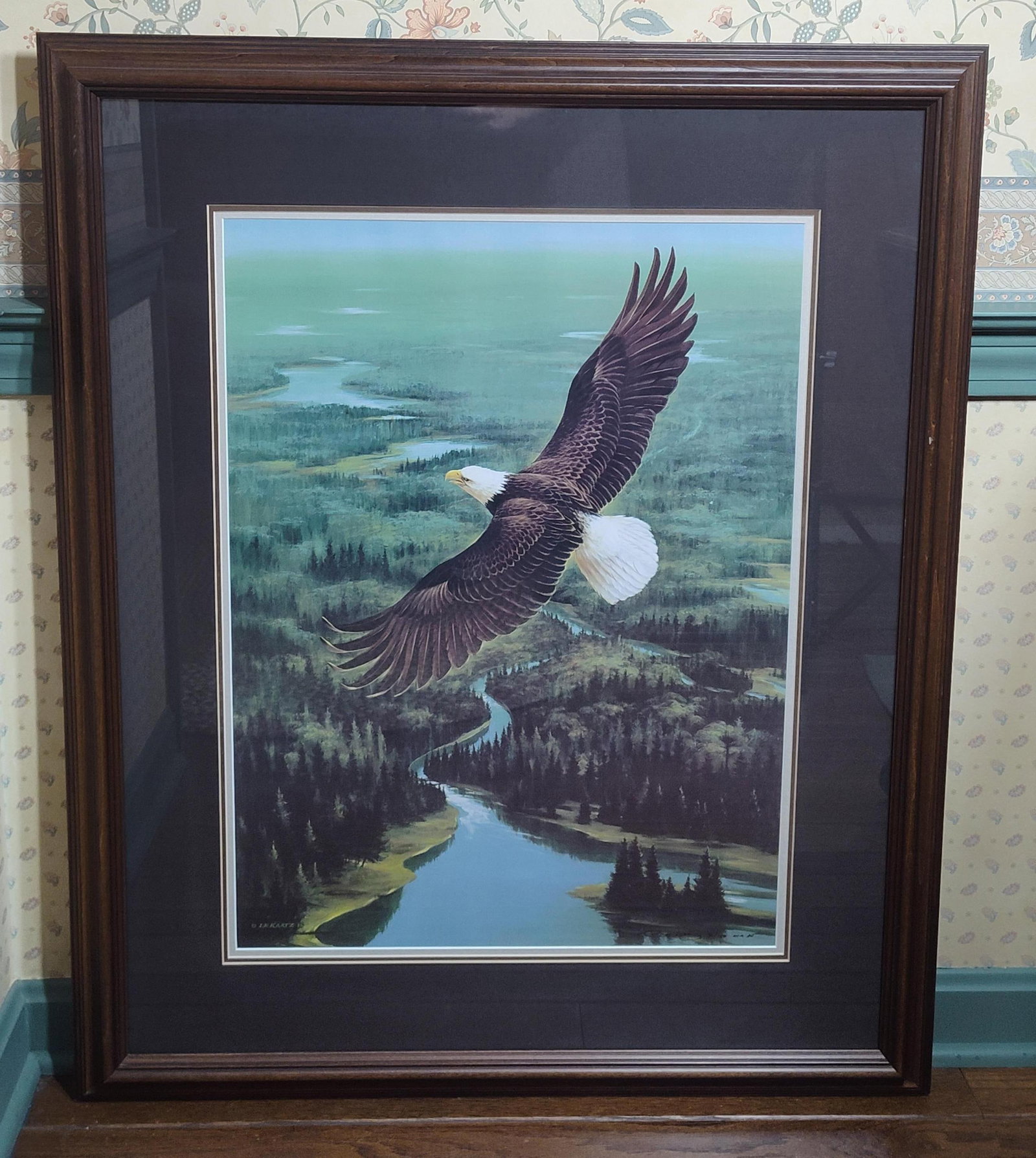 Lynn R. Kaatz Bald Eagle Offset Lithograph 30/150: This work is a limited edition offset lithograph by wildlife artist Lynn R. Kaatz (American, 20th century). The print depicts a bald eagle soaring above a coniferous forest landscape with a winding ri