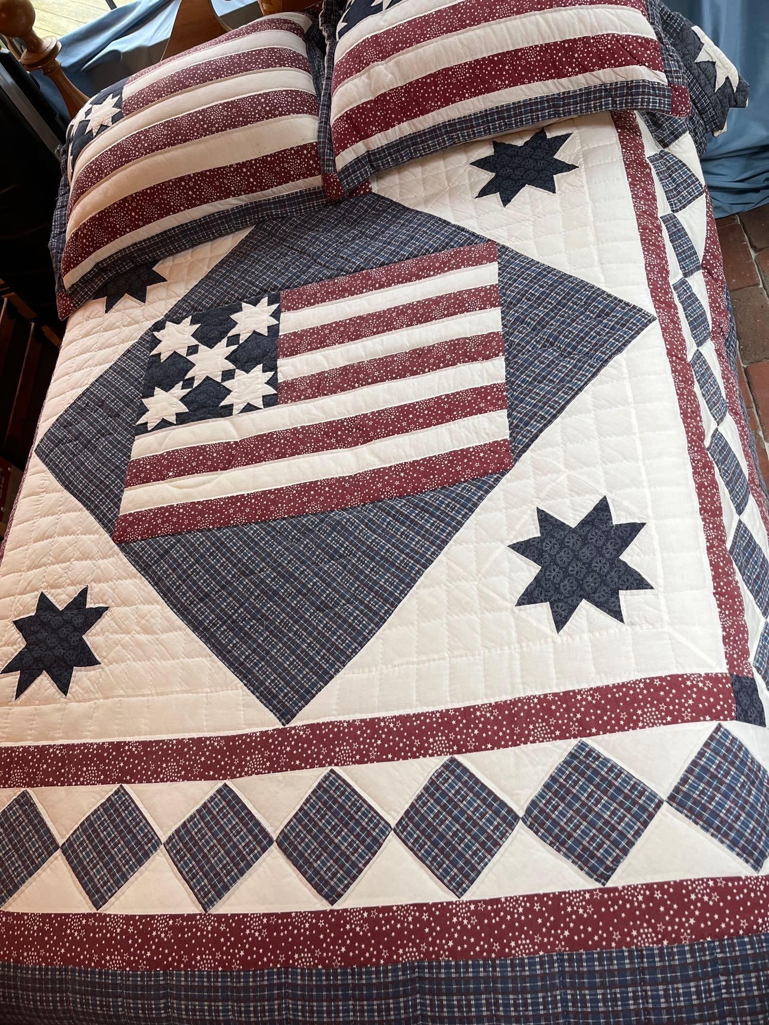 Patriotic American Flag & Star Motif Quilt Set: A three piece patriotic-themed bedding set comprising one quilt and two matching pillow shams. The design centers on a pieced American flag motif, featuring a navy blue canton with white stars