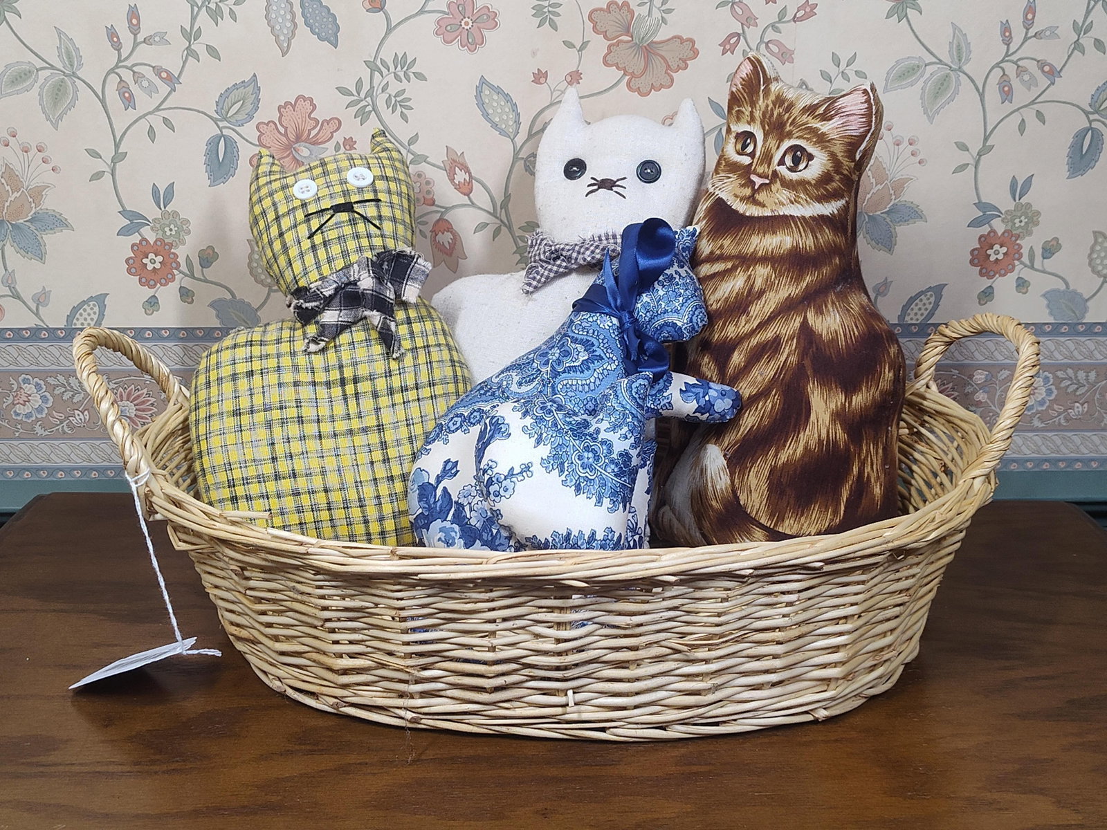 Folk Art Style Stuffed Cloth Cats in Wicker Basket: This lot features a collection of four folk art style stuffed cat figures presented within a light-colored woven wicker basket. The group includes one cat constructed from yellow and black plaid fabri